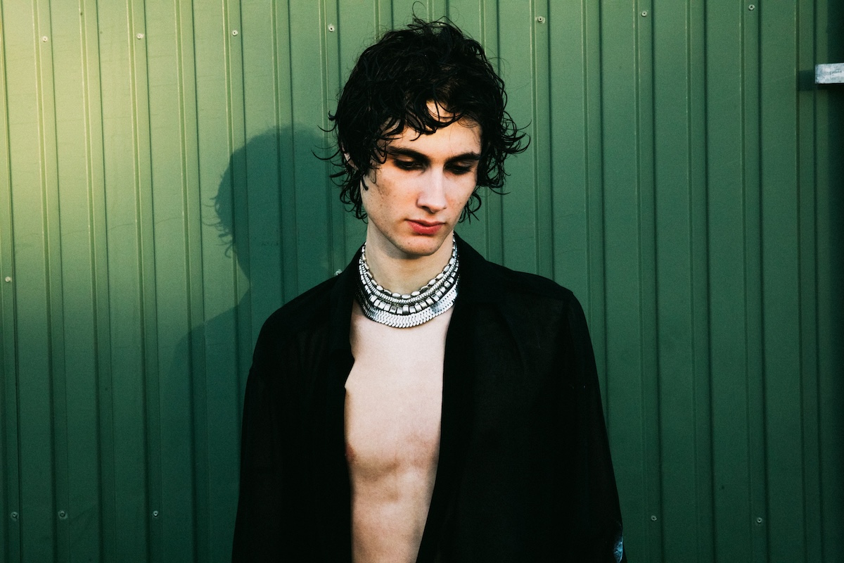 Promotional portrait of musician sombr (Shane Boose) wearing silver layered necklaces and an open black shirt against a green corrugated wall.